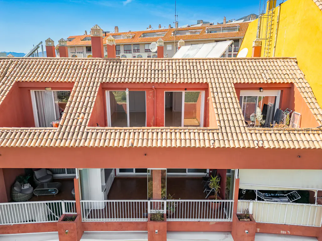Exterior view of a terracotta-colored building with a tiled roof and balconies with white railings. Three dormer windows are visible on the top floor.