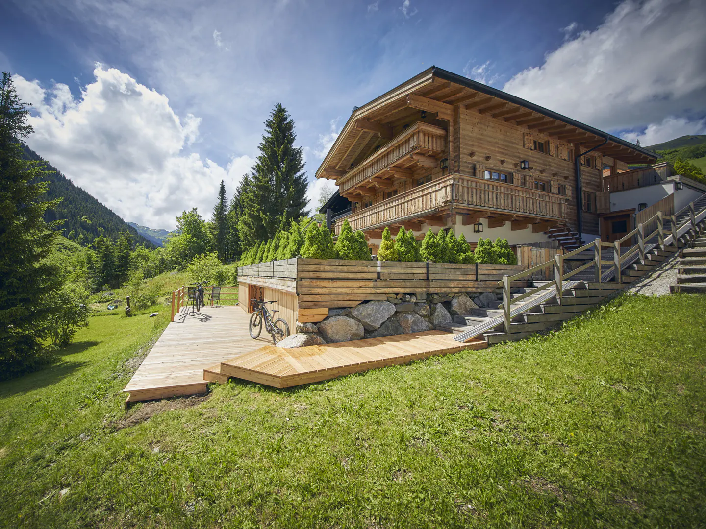 Exterior view of a wooden chalet with balconies, a deck, and a bike, set against a backdrop of green hills and a blue sky with clouds.