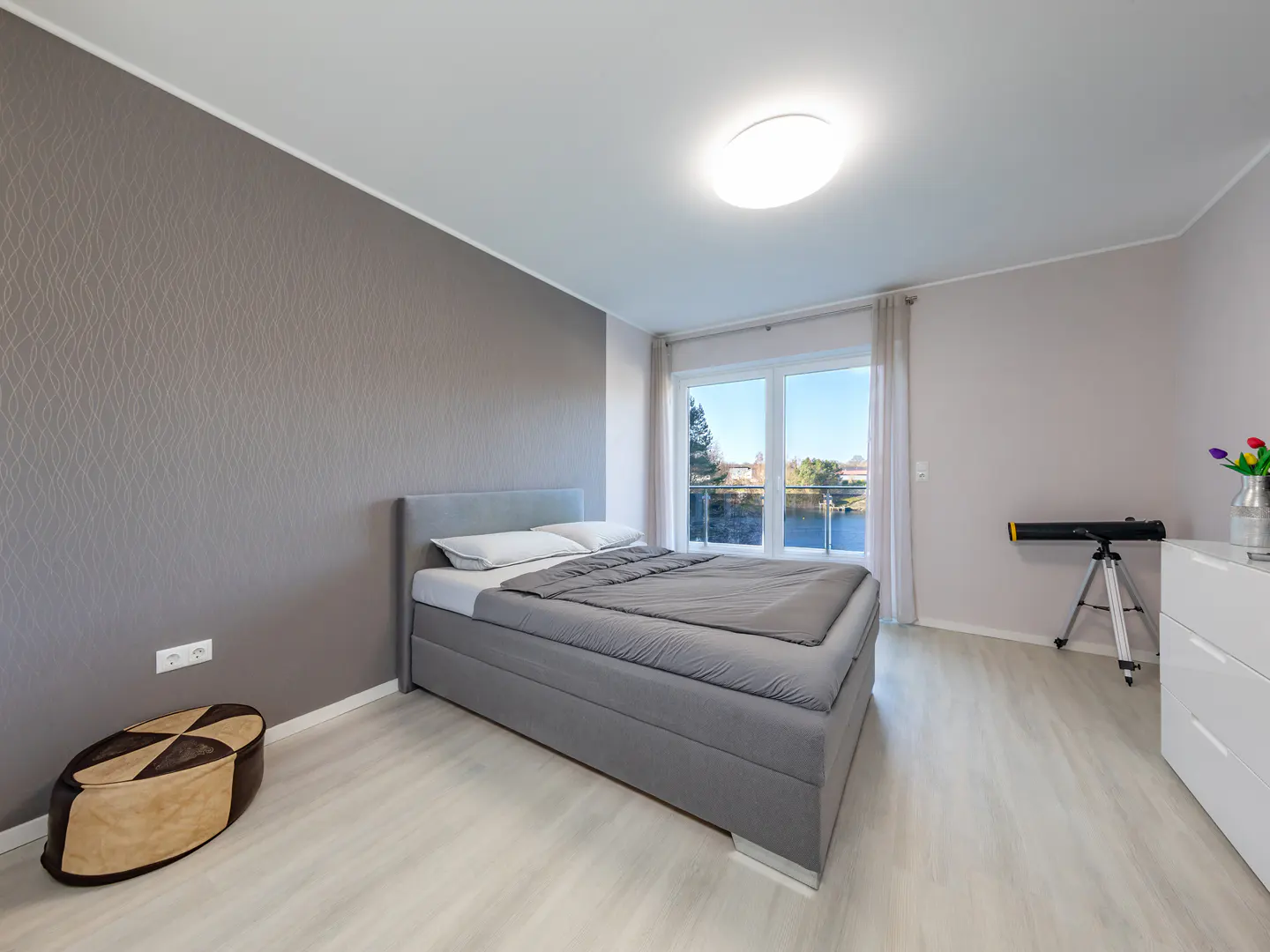 A modern bedroom with a gray bed, light wood floors, and a large window with a view. A telescope stands near a white dresser.
