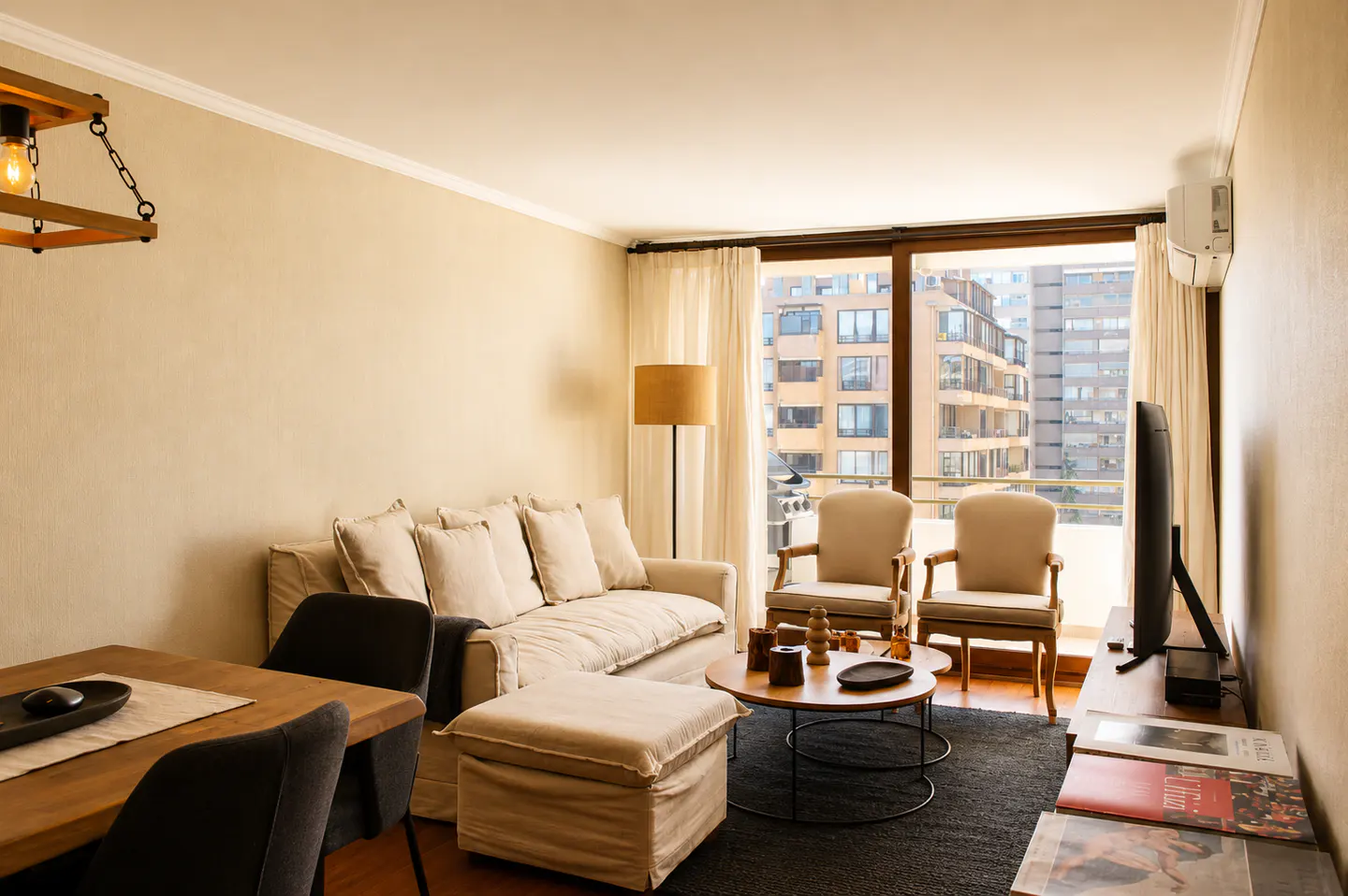 Living room with beige sofa, chairs, and ottoman. A round coffee table sits on a dark rug. Large windows overlook city buildings.