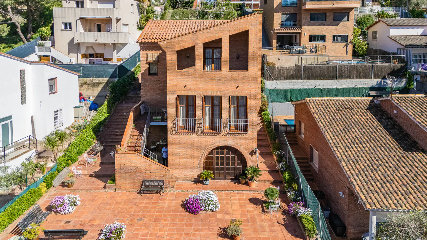 Aerial view of a three-story brick house with a red tile roof and a large arched doorway. The house has a tiled patio with flowers and benches.