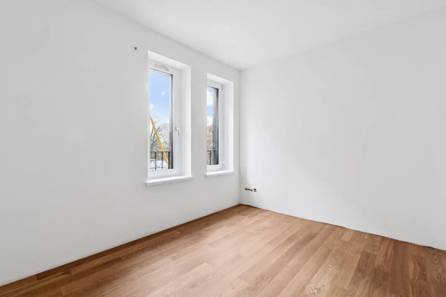 Bright, empty room with white walls, wood floors, and two windows showing a balcony and trees.