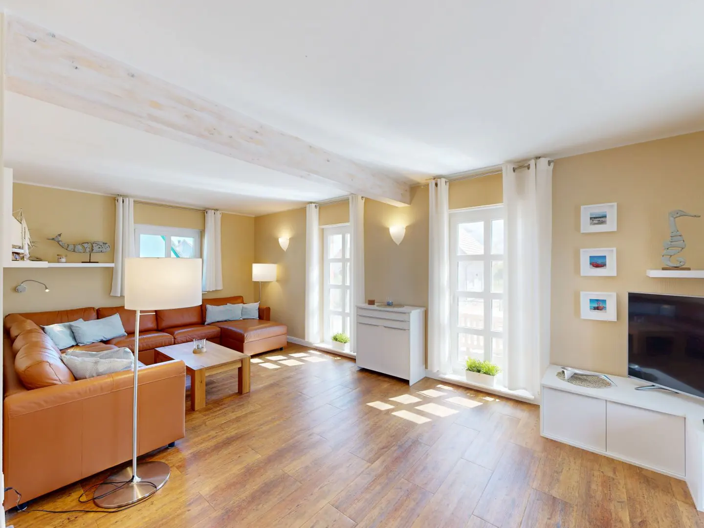 Bright living room with a brown leather sectional sofa, wood floors, white walls and trim, and large windows with white curtains.