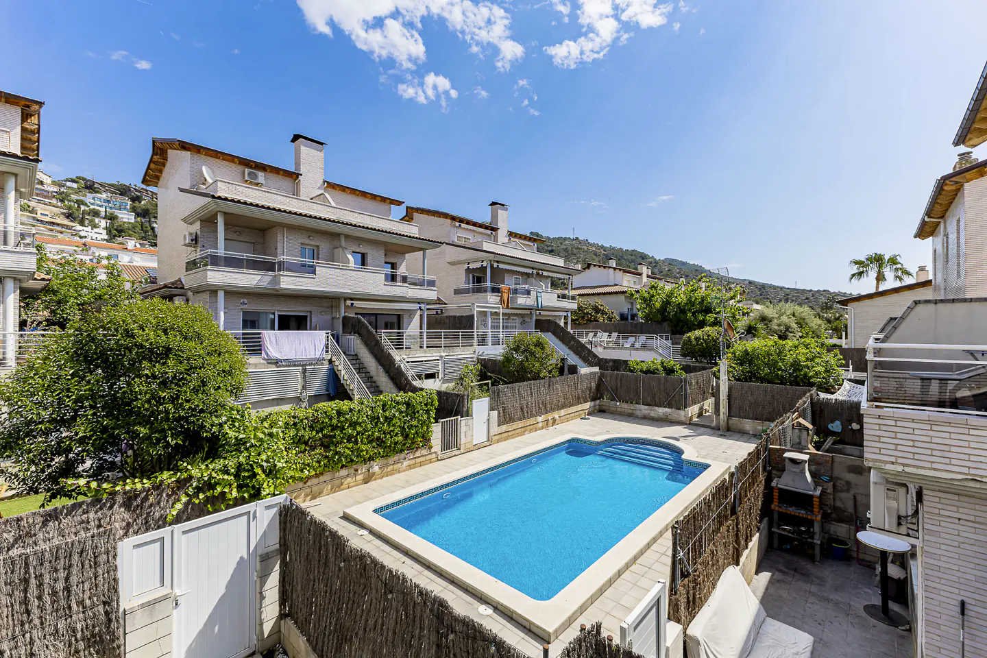 View of a blue swimming pool surrounded by townhouses with balconies and stairs, under a blue sky.