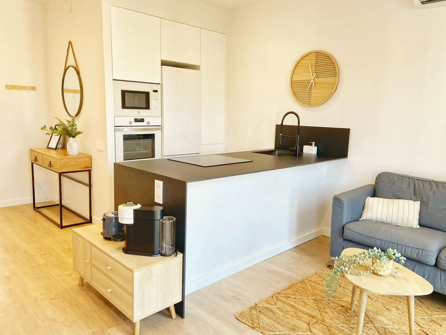 A bright, modern apartment interior with a kitchen island, gray sofa, and wooden accents. Appliances and decor create a cozy, stylish living space.