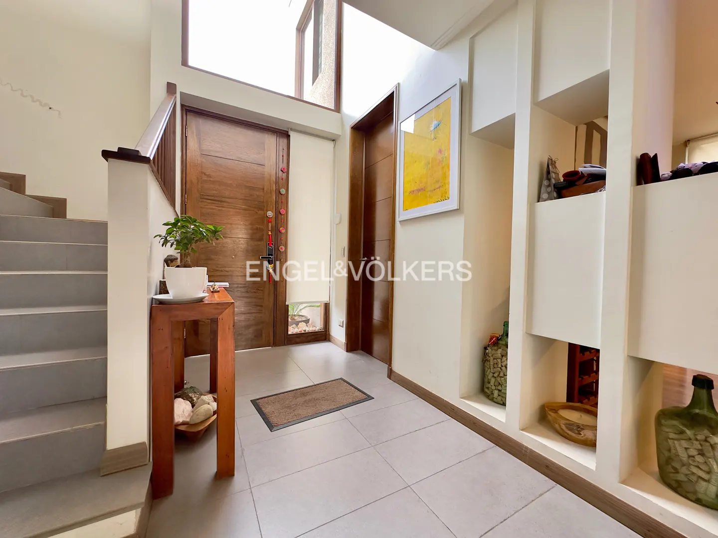 Bright entryway with a wooden door, staircase, tiled floor, and built-in shelving featuring decorative items and art.