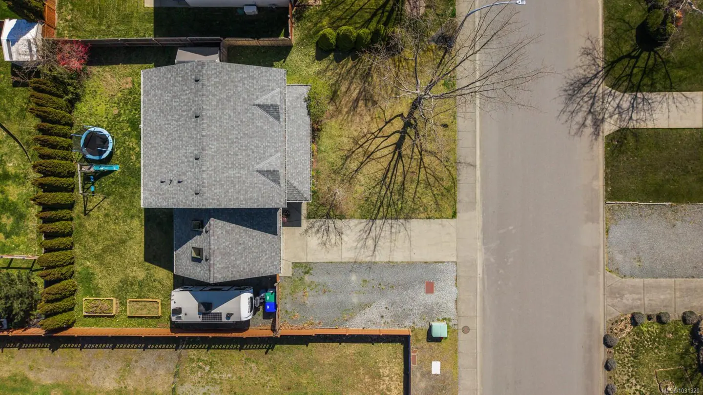 Aerial view of a house with a gray roof, green lawn, trampoline, RV, and a tree with bare branches.