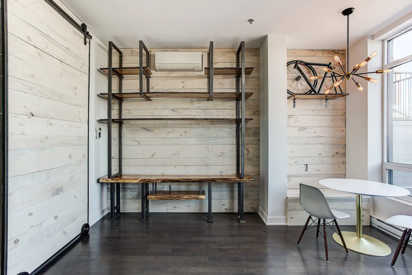 Modern interior with white wood paneling, dark wood floors, and a metal and wood shelving unit. A white table and chairs sit near a window.