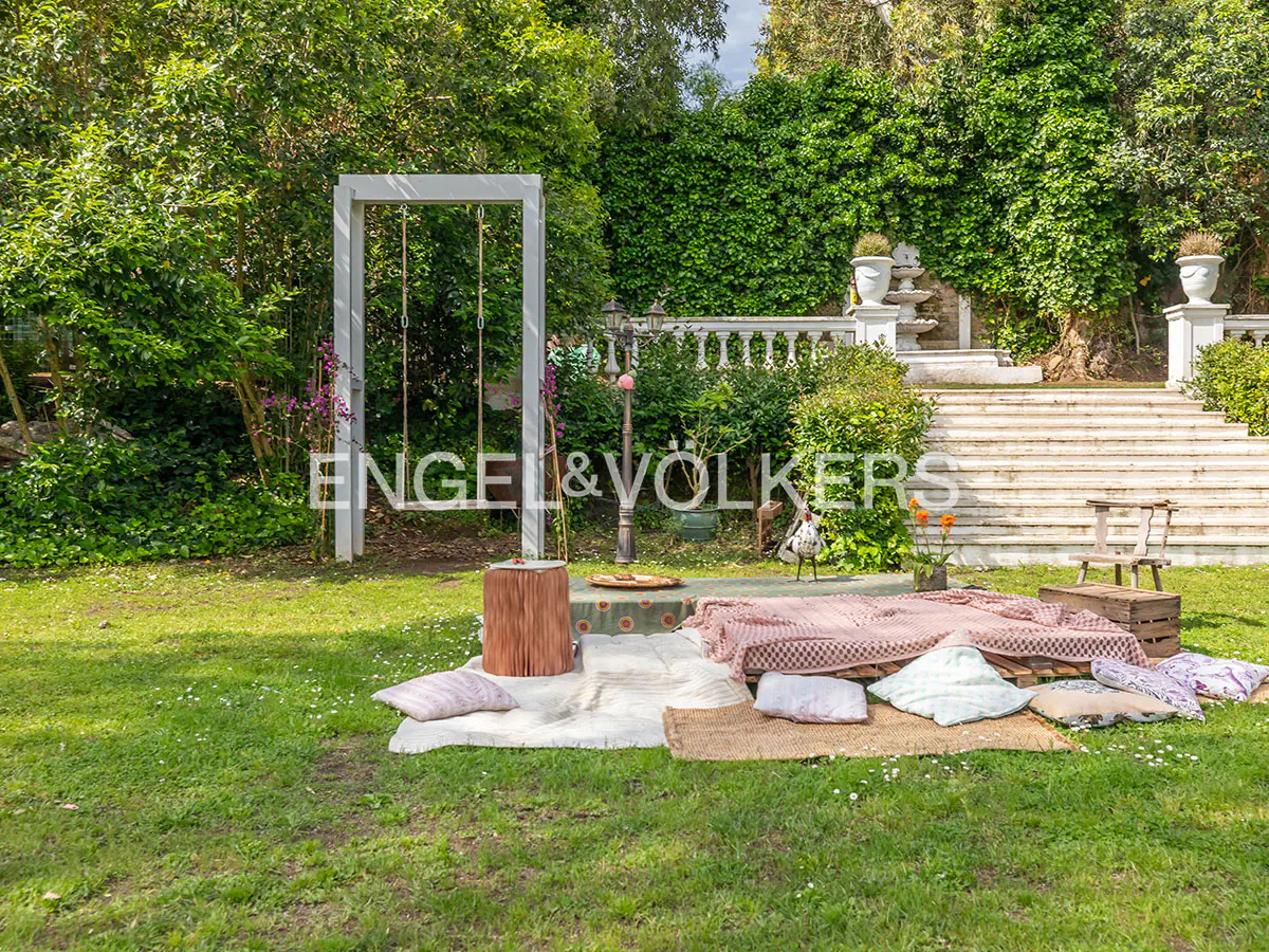 Lush green lawn with picnic setup: blankets, pillows, and a wooden swing. Stone stairs and a balustrade are in the background.