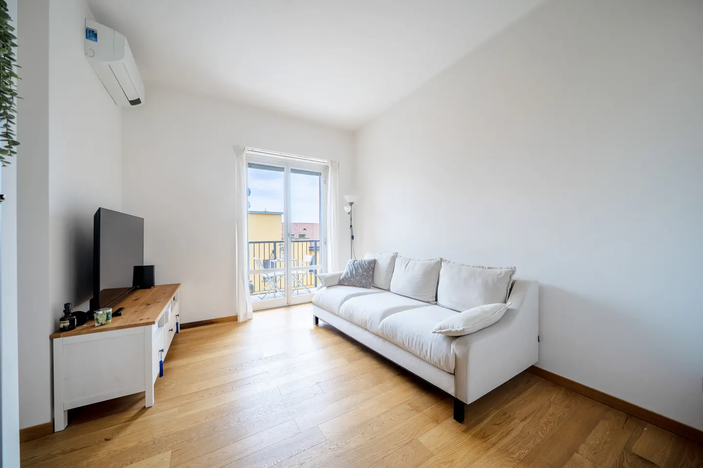 Bright living room with white walls, wood floors, and a white sofa. Balcony doors let in natural light. TV on a white cabinet.