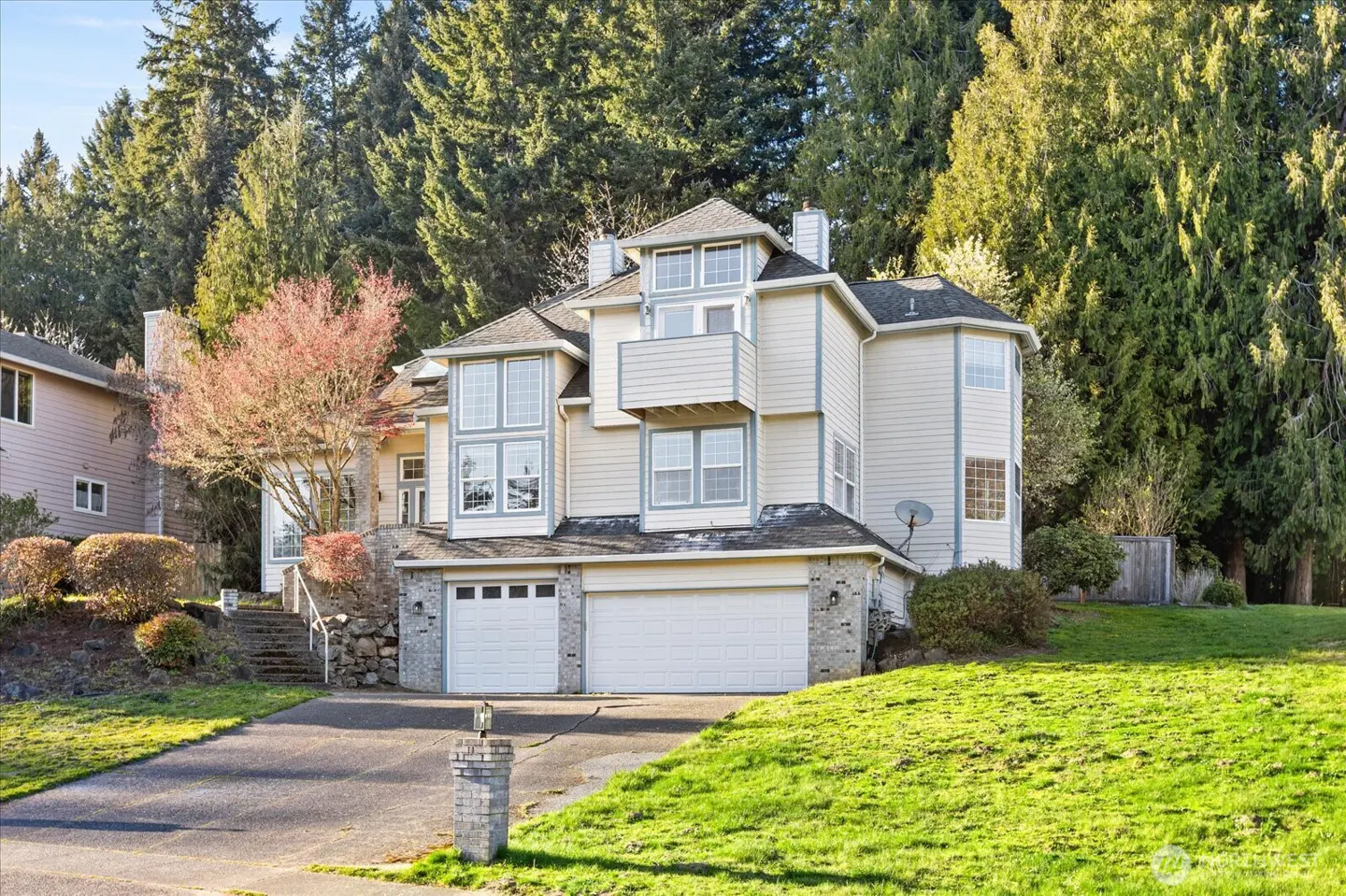 A multi-story beige house with a gray roof and white garage doors sits on a green lawn with tall trees in the background.