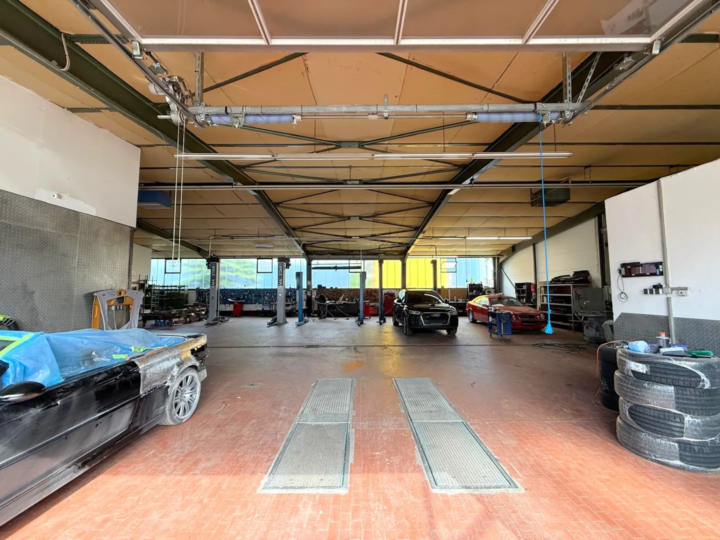 A wide shot of a large auto repair shop with several cars, lifts, and tools. The floor is red tile and the ceiling is high with metal beams.