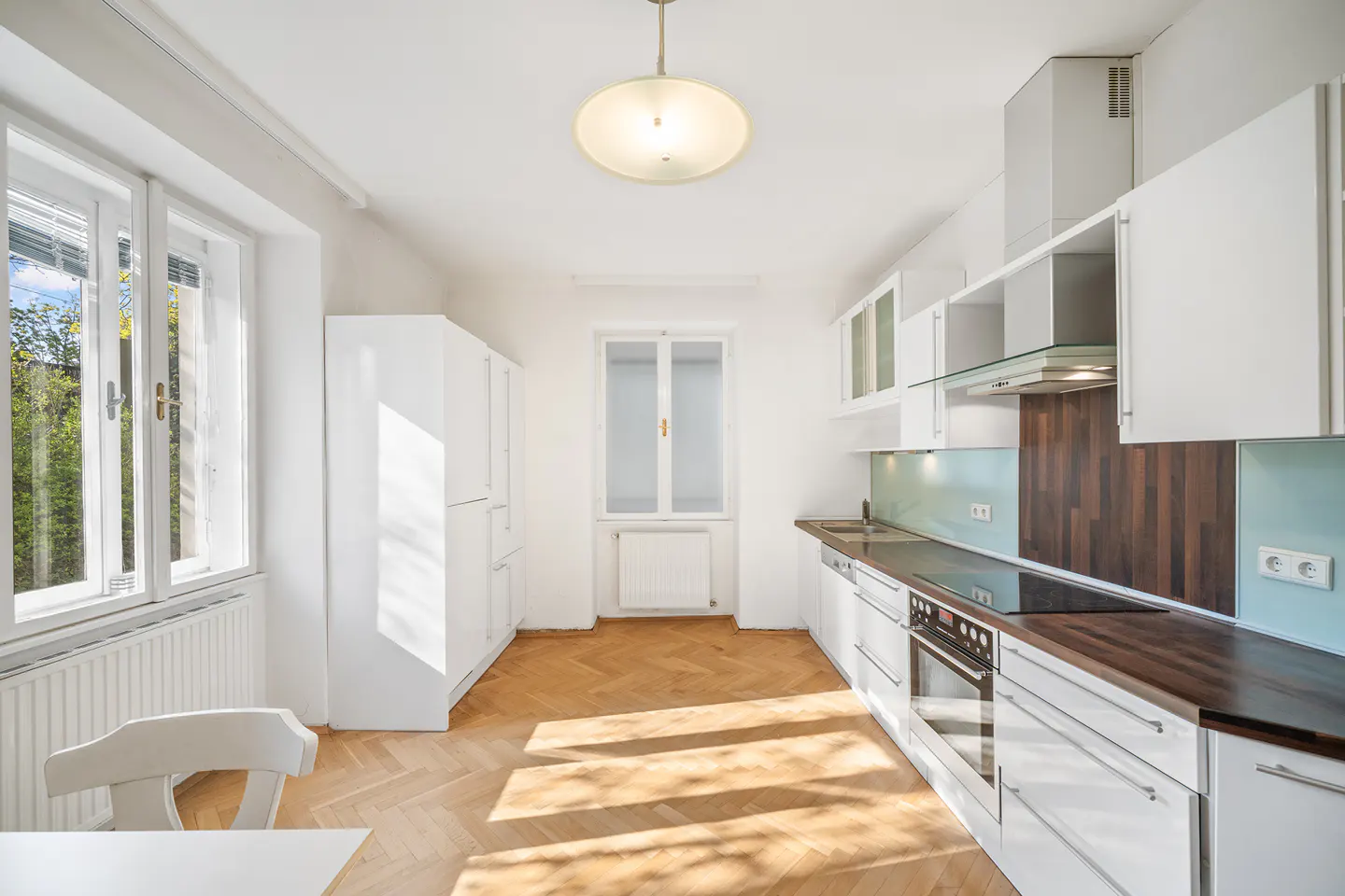 Bright, empty kitchen with white cabinets, wood countertops, and herringbone floors. Sunlight streams through the window.