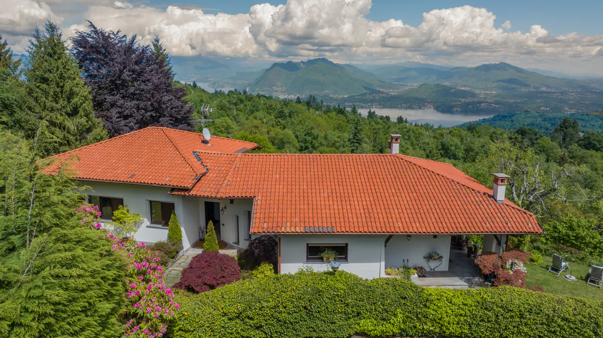A white house with a red tile roof is surrounded by green trees and bushes, with mountains and a lake in the background.