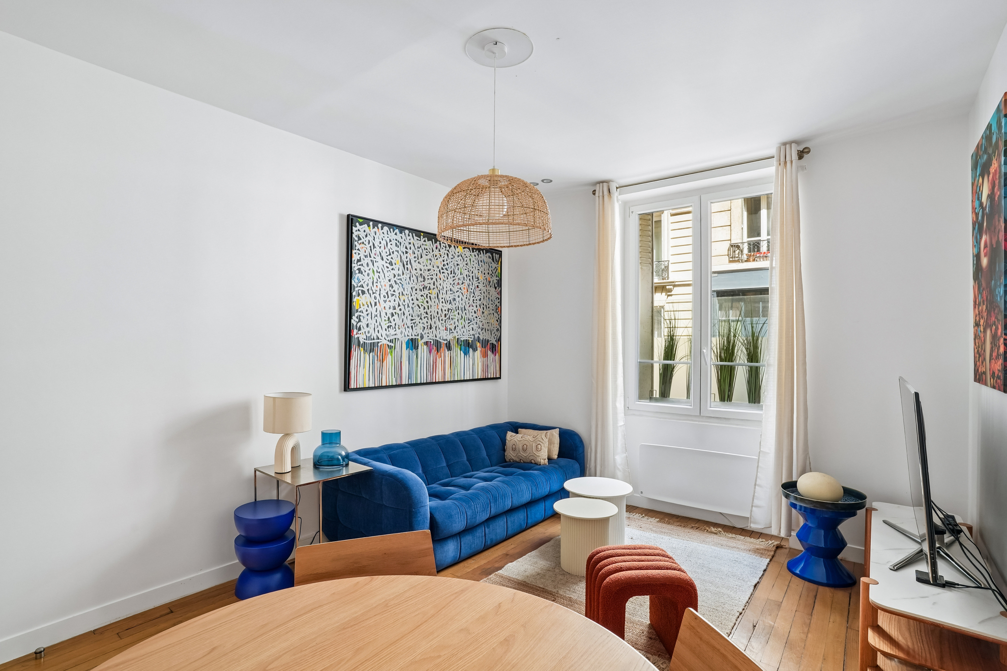 Bright living room with a blue velvet sofa, wooden dining table, modern art, and a woven pendant light by a window. [AI-generated text]