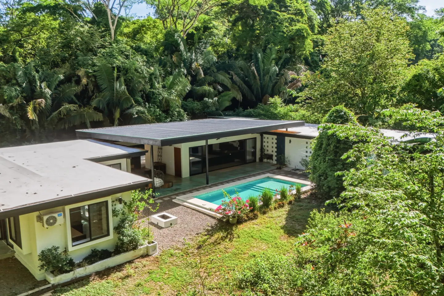 Aerial view of a modern, white house with a black roof, a turquoise pool, and lush green trees in the background.