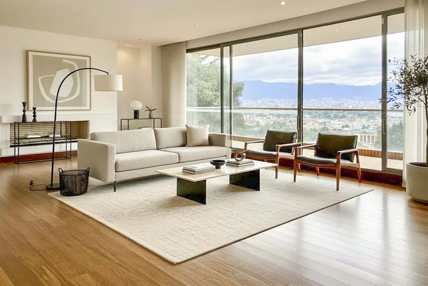 Modern living room with beige sofa, two leather armchairs, coffee table, and floor-to-ceiling windows overlooking a city.
