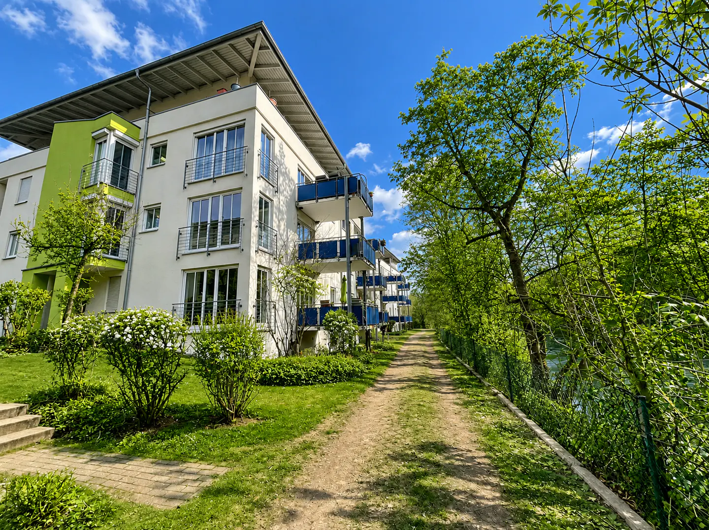Modern white apartment building with blue balconies, green accent wall, and a path leading alongside it, under a bright blue sky.