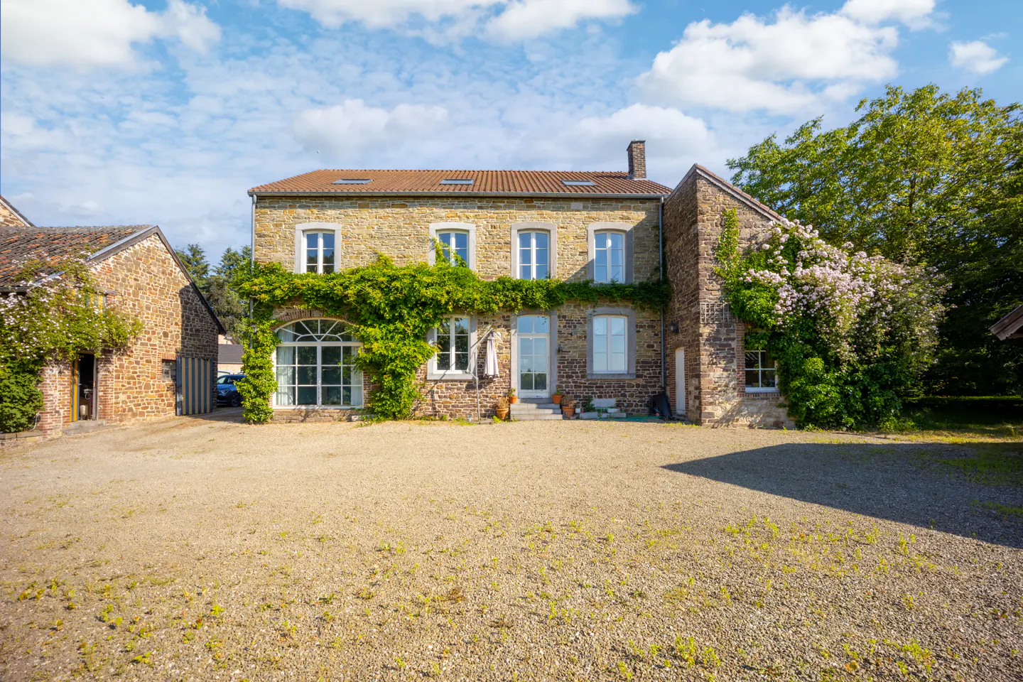 A two-story stone house with a gravel driveway, climbing vines, and a blue sky with clouds.