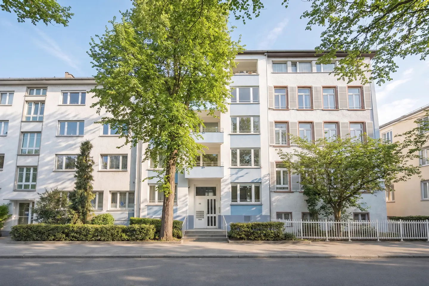 Exterior of a light blue and white apartment building with green trees and bushes in front.