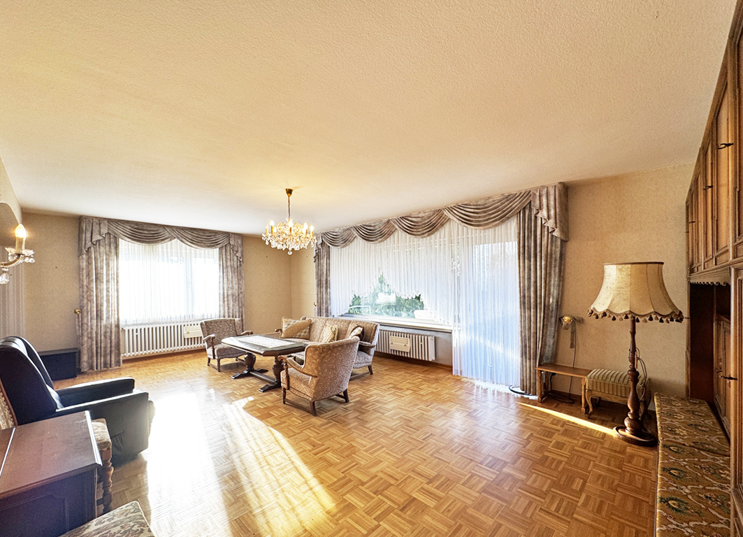A spacious living room with parquet flooring, a chandelier, and large windows with decorative curtains. Furniture includes armchairs, a sofa, and a lamp.