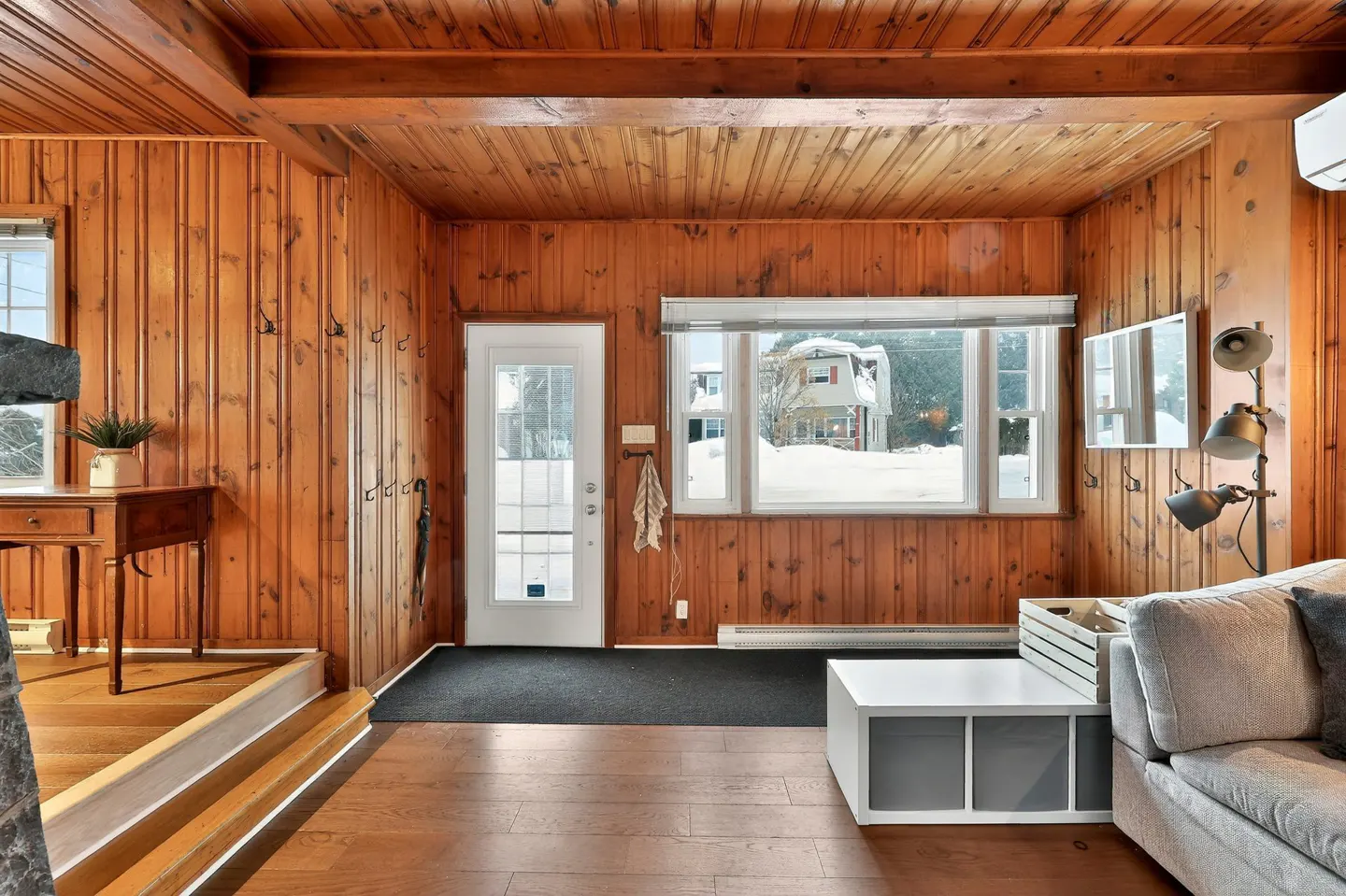 Interior view of a wood-paneled room with a white door, window with snowy view, and a gray sofa.