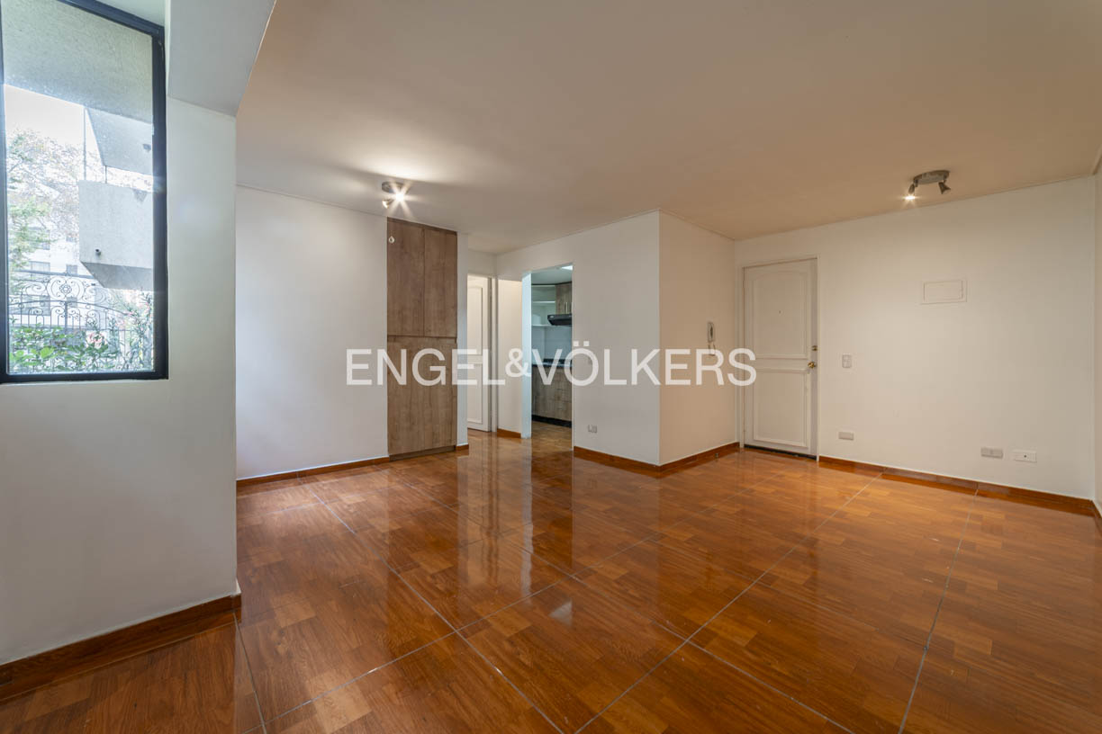 Bright, empty room with glossy wood floors, white walls, and a large window. A kitchen is visible through an open doorway. Engel & Völkers logo is centered.