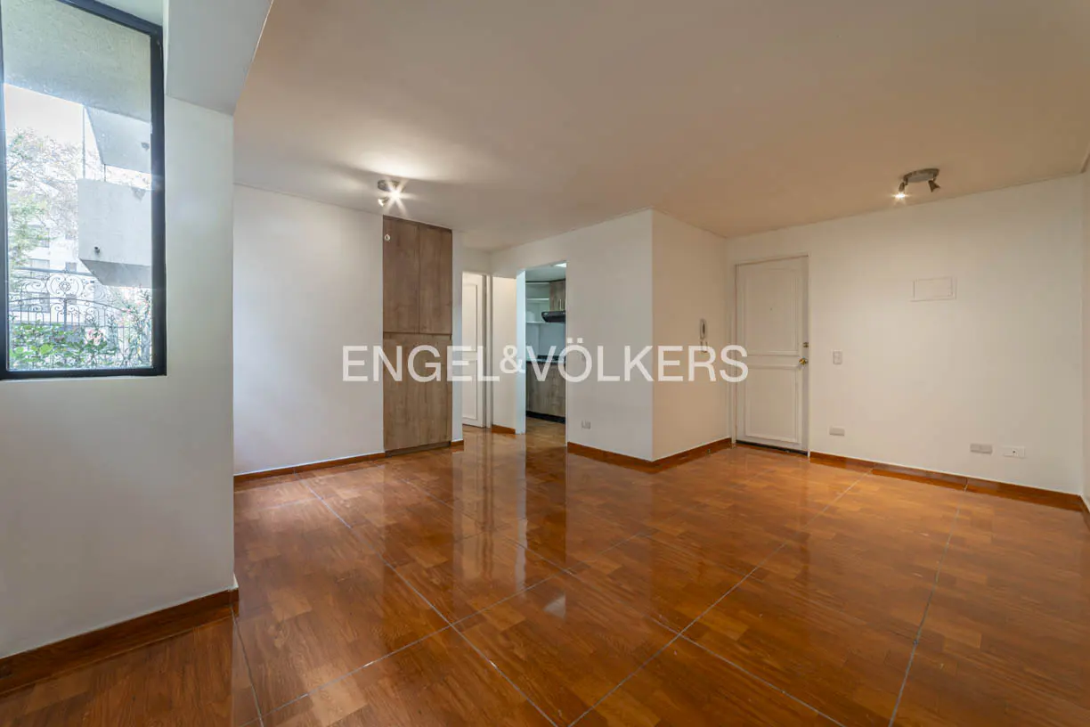Bright, empty room with glossy wood floors, white walls, and a large window. A kitchen is visible through an open doorway. Engel & Völkers logo is centered.