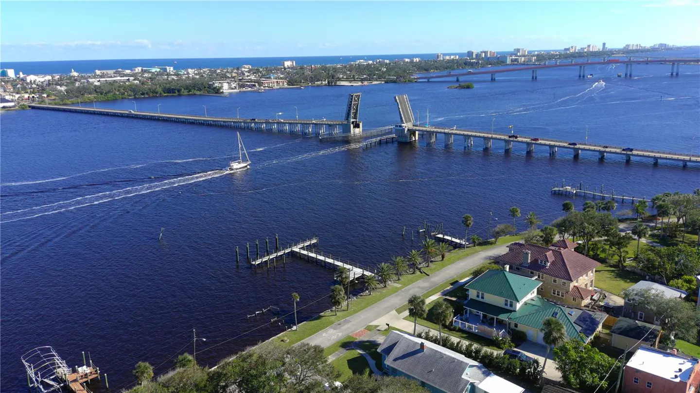 Aerial view of a blue river with a sailboat, a drawbridge, and waterfront homes on a sunny day.