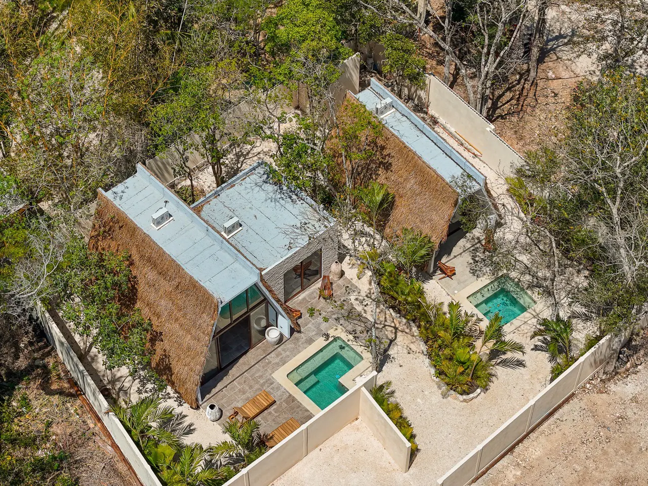 Aerial view of two modern homes with thatched roofs, small pools, and surrounded by lush greenery and a white perimeter wall.