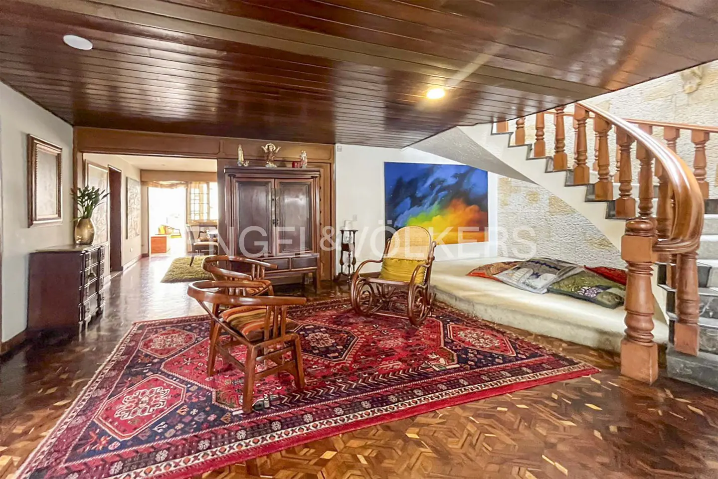 A living room with wooden floors, a red rug, two chairs, a rocking chair, and a staircase. A painting hangs on the wall.