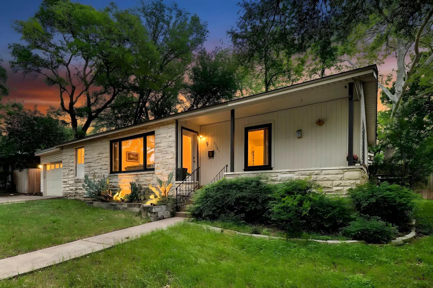 Exterior view of a single-story home with stone and white siding, black window frames, and a green lawn at dusk.