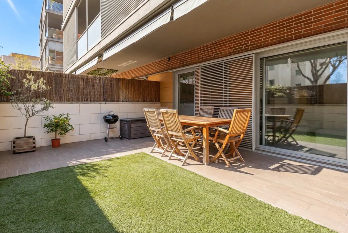 Outdoor patio with a wooden table and chairs, a grill, and a small lawn. Sliding glass doors lead into the building.
