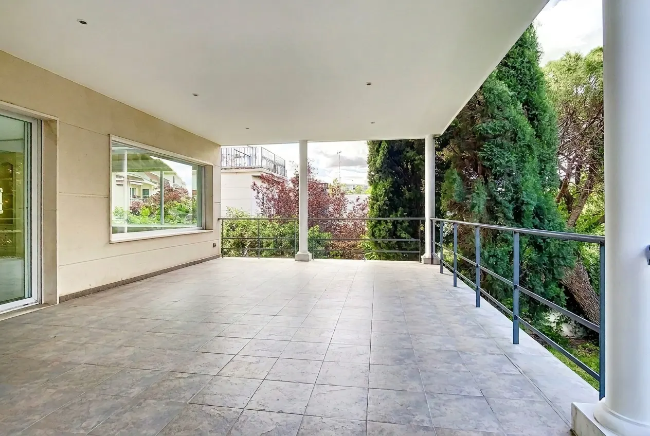 Covered patio with gray tiled floor, white columns, and black metal railing overlooking trees and a neighboring house.