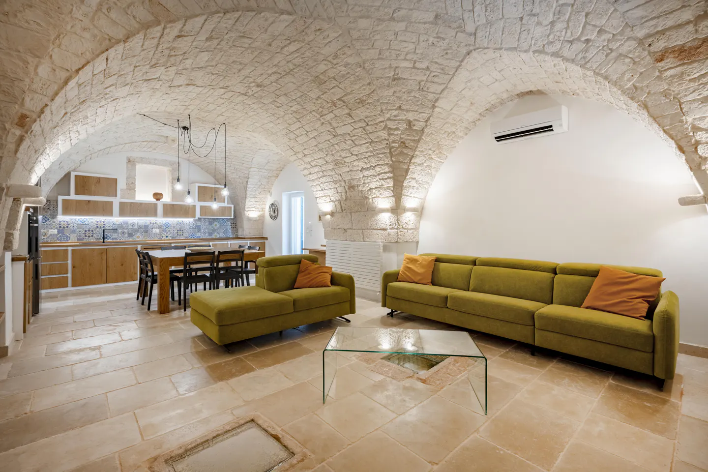 Living room with stone vaulted ceiling, olive sofas with orange pillows, glass table, and kitchen in the background.