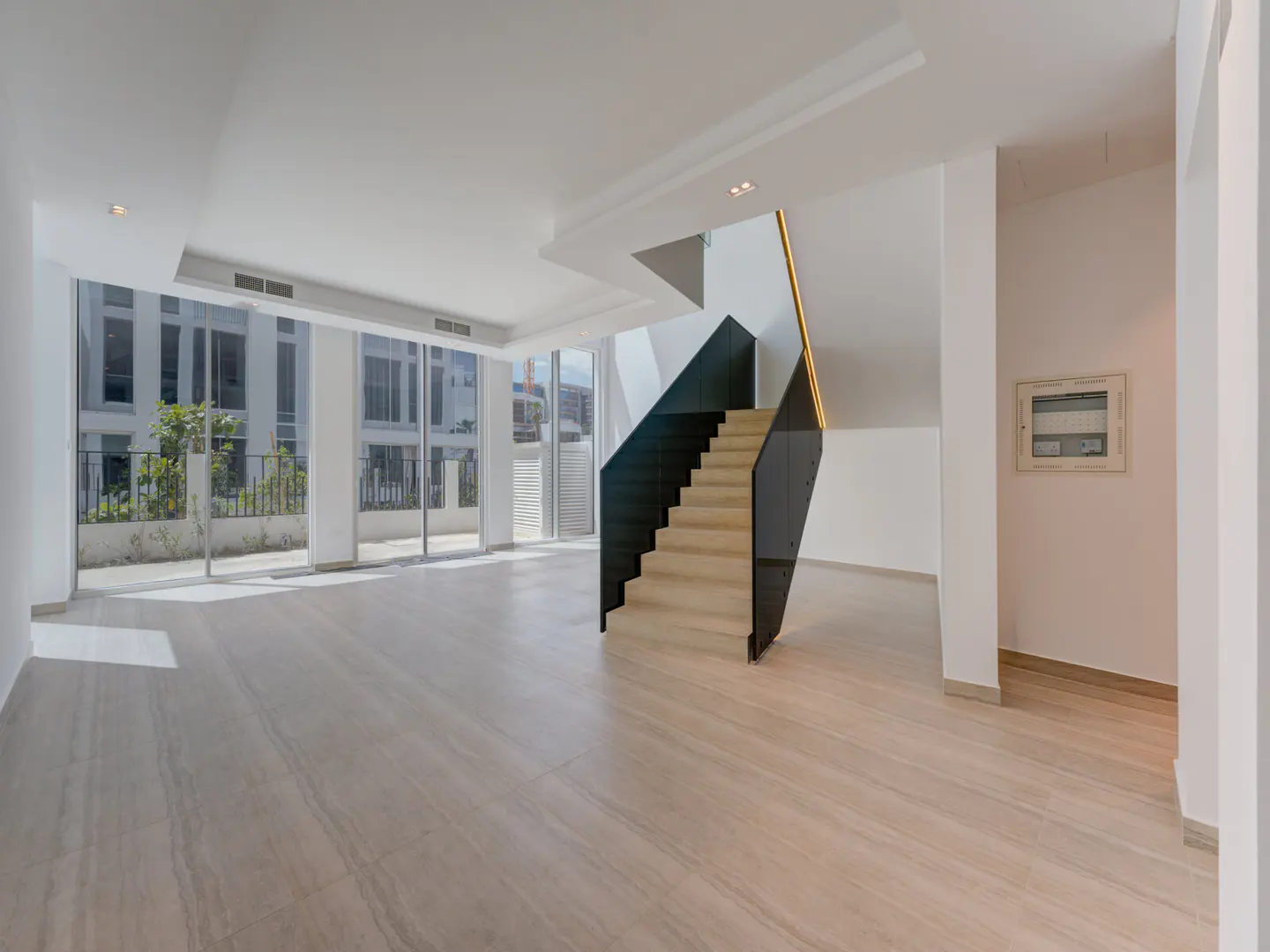 Bright, empty room with light wood floors, white walls, and a modern staircase with black accents. Large windows offer a view of a building and greenery.