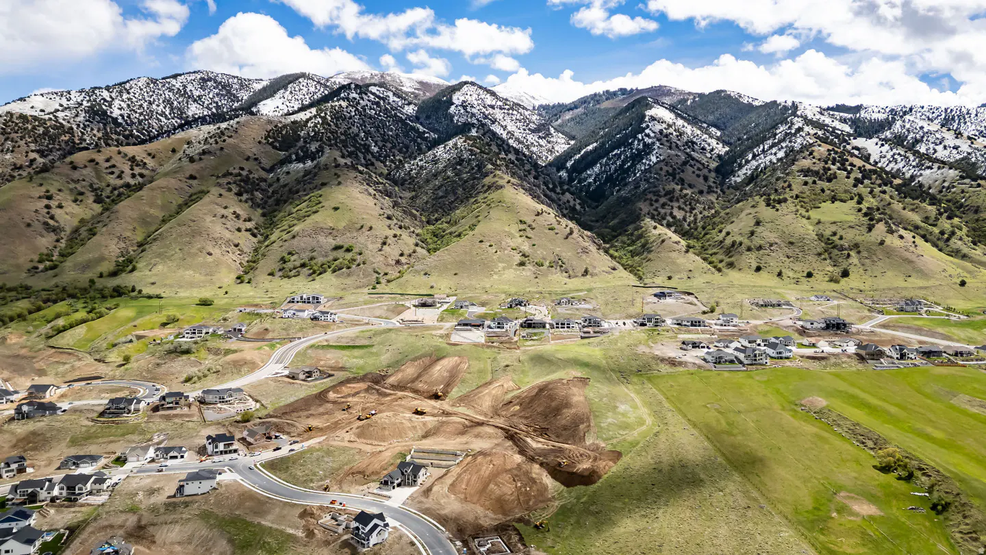 Aerial view of a housing development at the base of snow-capped mountains under a blue sky with white clouds.
