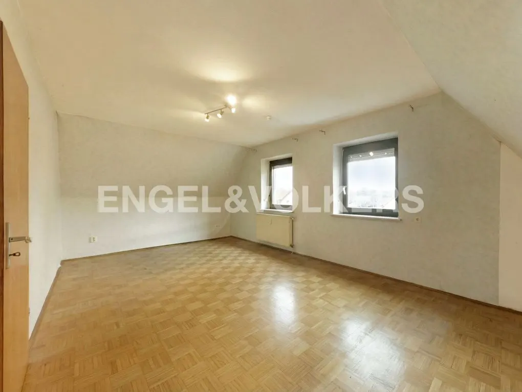 Bright, empty attic room with parquet floor, white walls, two windows, and a door on the left.