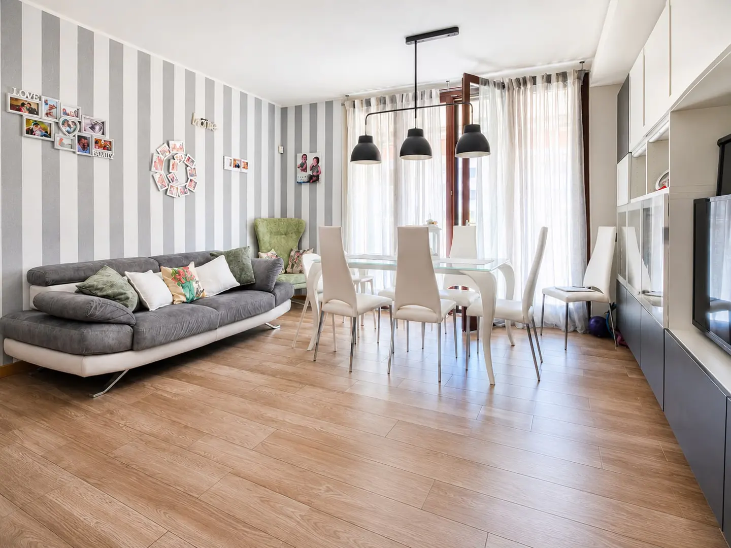 Modern living and dining room with grey striped walls, a grey sofa, glass dining table, and white chairs on wood floors.