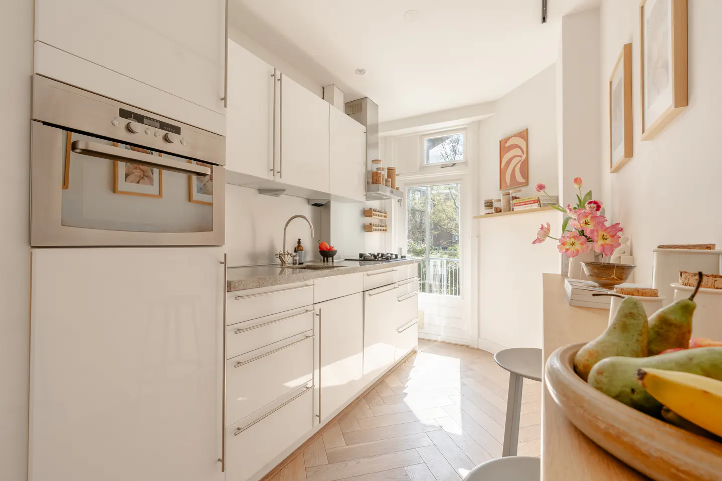 Bright, modern kitchen with white cabinets, stainless steel oven, and herringbone wood floors. Fruit bowl and flowers add color.