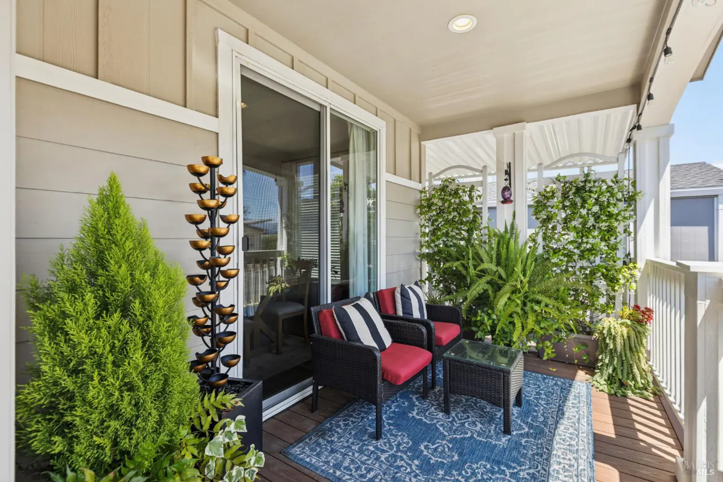 A cozy outdoor patio with two black wicker chairs with red cushions, a blue rug, and lush greenery. A water fountain adds a tranquil touch.