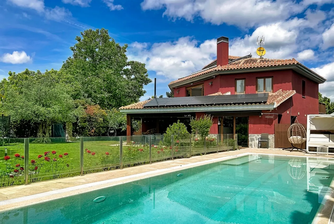 A red house with a pool, lawn, and trees under a blue sky with white clouds.