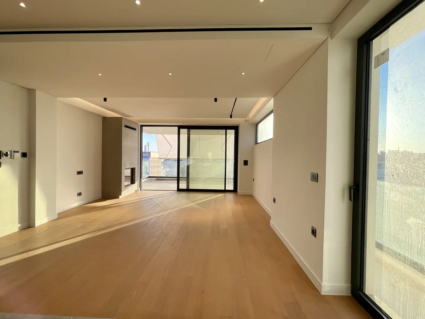 Empty living room with light wood floors, white walls, and black framed sliding glass doors to a balcony. Large window on the right.