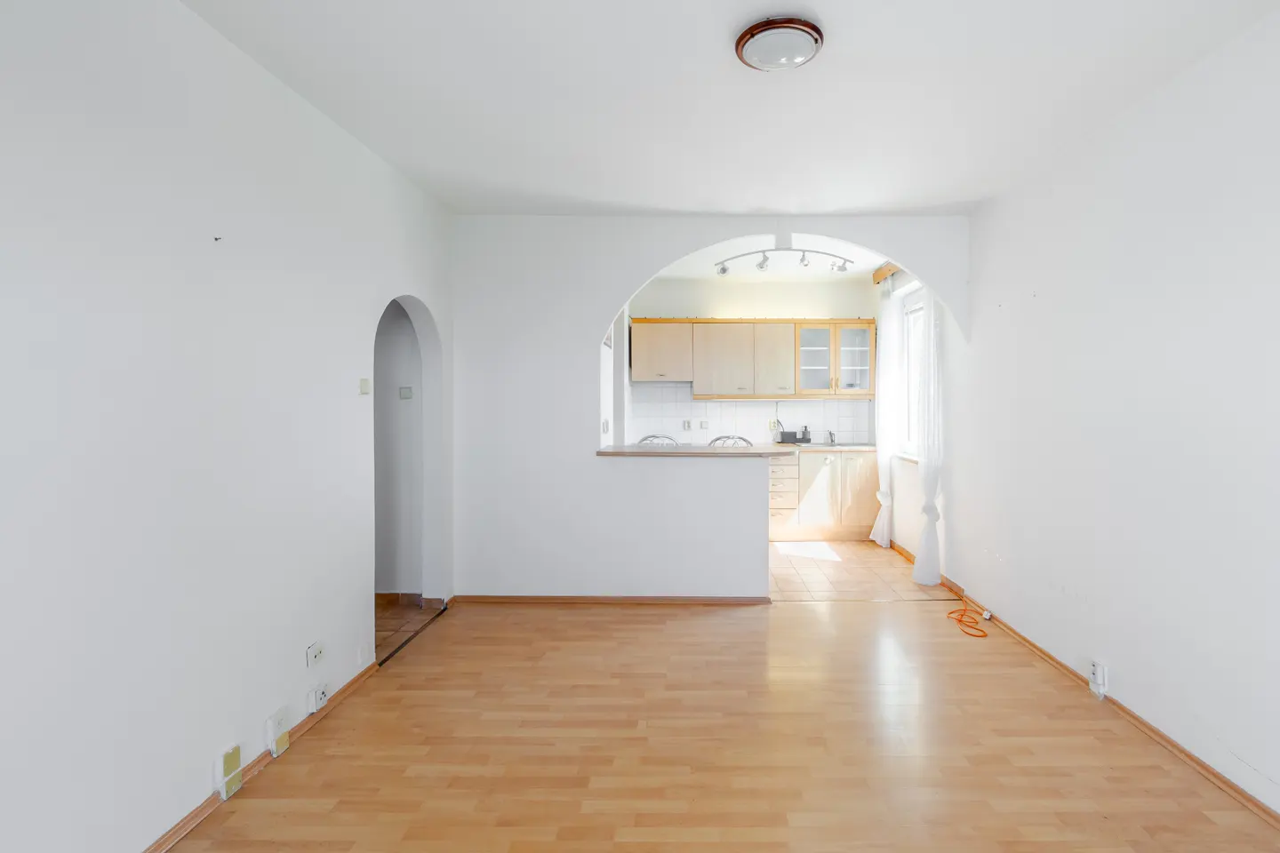 Bright, empty room with wood floors and white walls. An arched doorway leads to a kitchen with light wood cabinets.