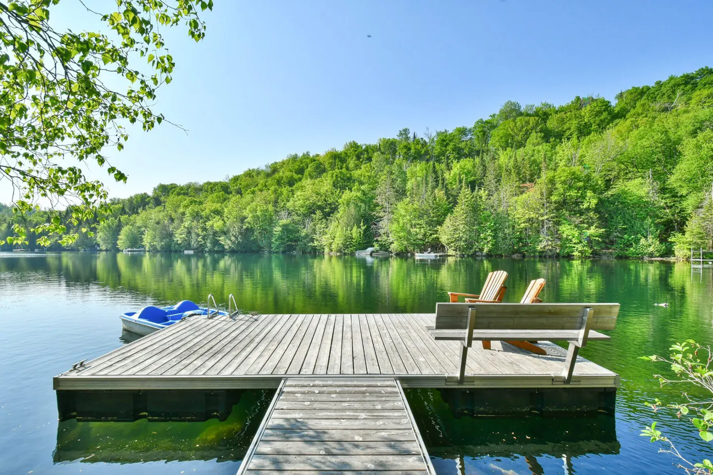 Wooden dock on a lake with chairs, bench, and paddle boat. Green trees line the shore under a blue sky.