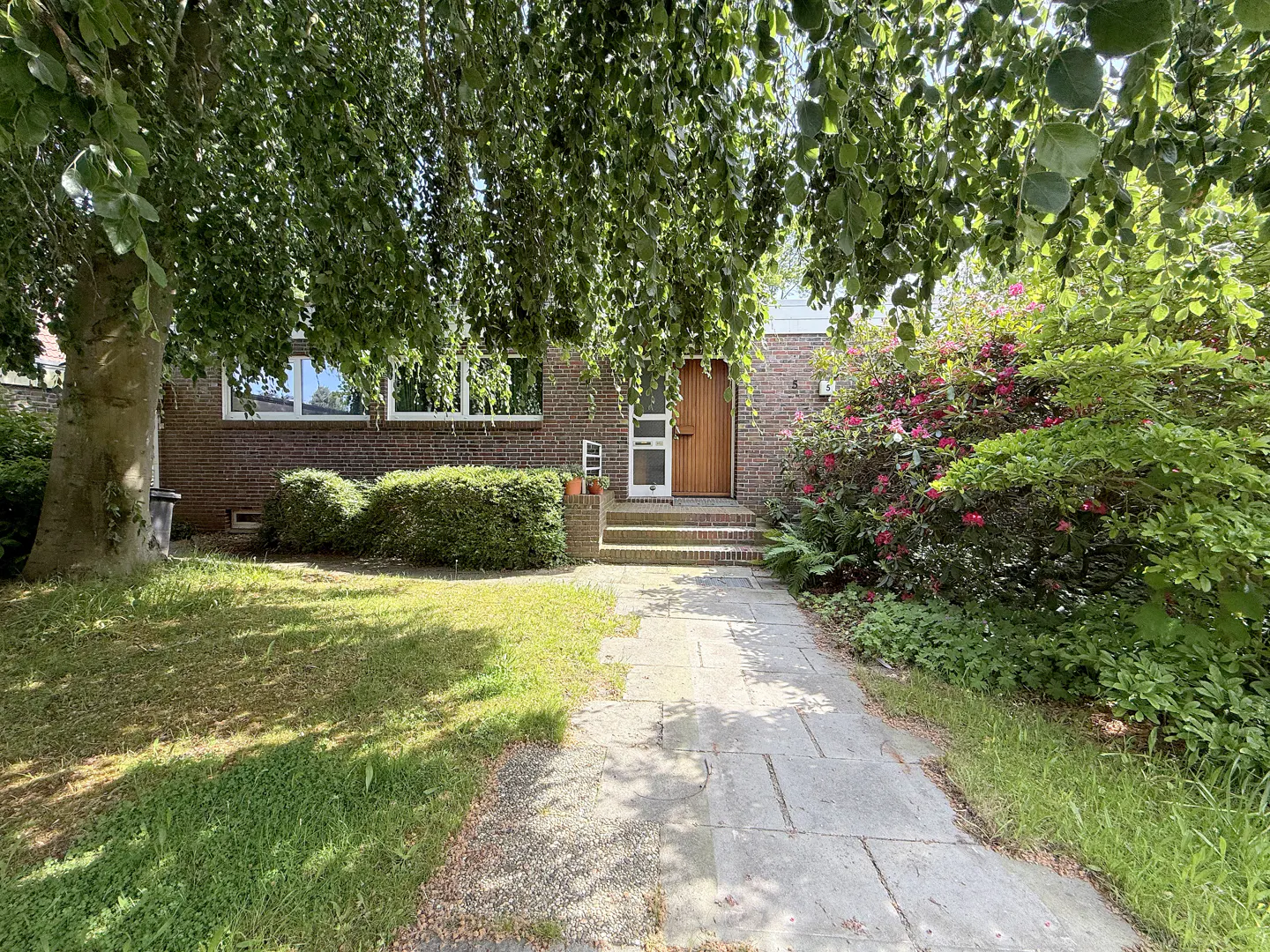 Brick house with a wooden door, steps, and a stone walkway, surrounded by green trees and bushes.
