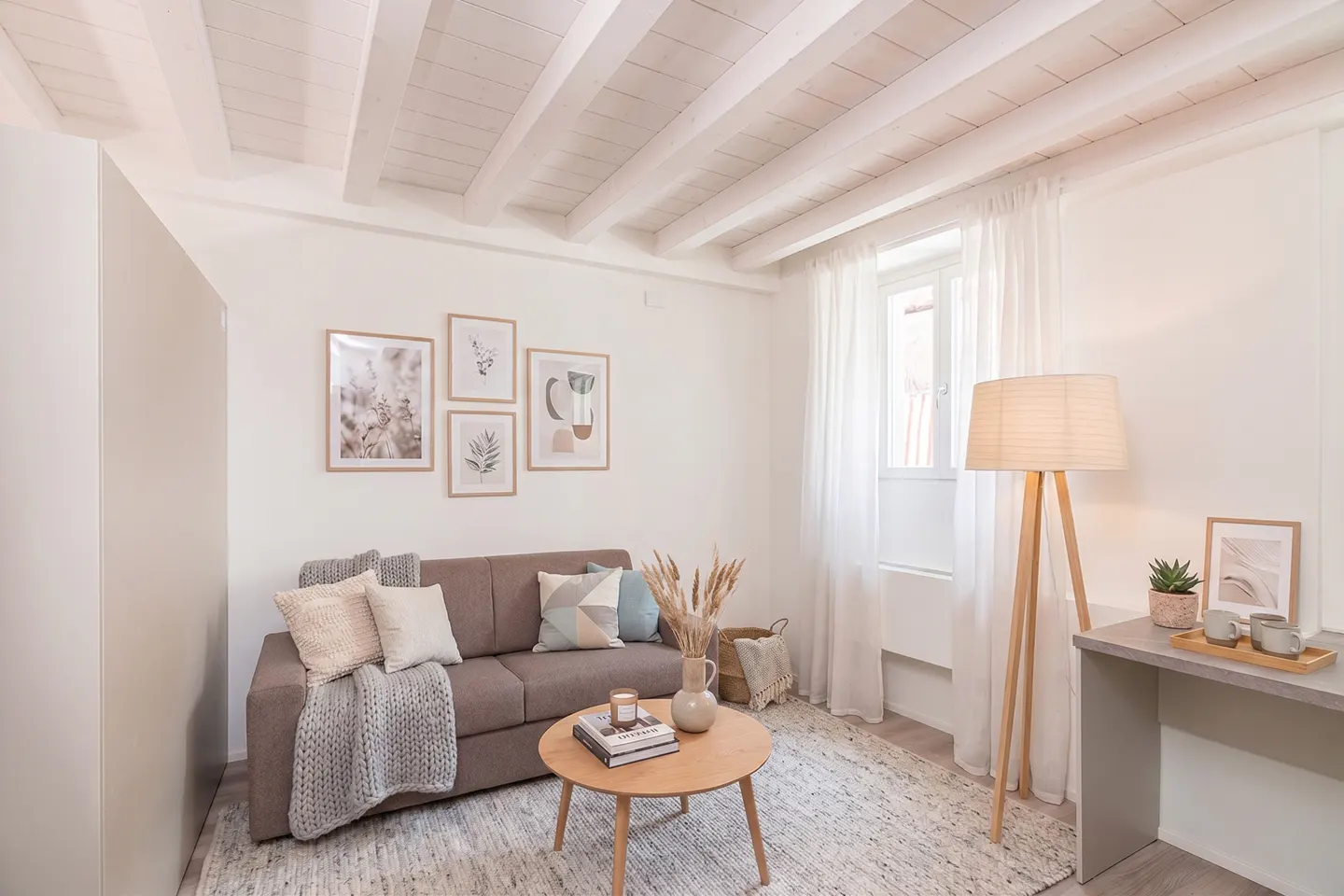 Cozy living room with a brown sofa, round wooden coffee table, floor lamp, and white beamed ceiling in a neutral palette.