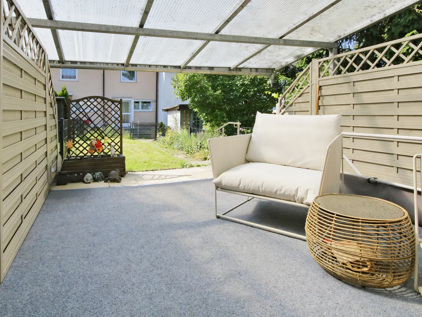 Outdoor patio with a white sofa, wicker table, and gray flooring under a clear roof. Fenced yard with greenery in the background.