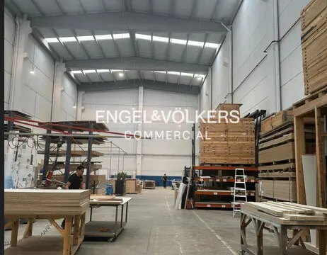 A large, bright warehouse interior with stacks of wood, tables, and an Engel & Völkers Commercial logo in the background.