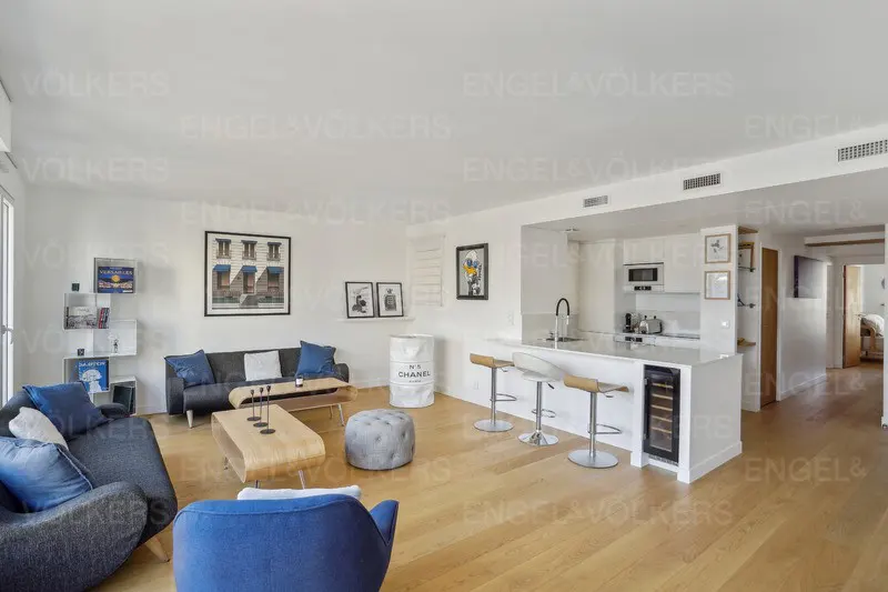 Bright, open-plan living space with wood floors, white walls, and blue accents. Kitchen island with bar stools and modern appliances. Artwork on walls.